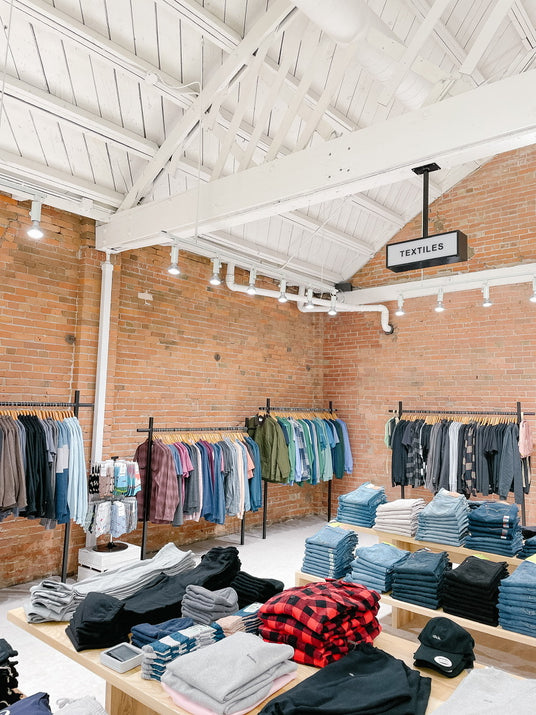A well-lit retail store with exposed brick walls, filled with racks of clothing and tables with shirts & pants folded on them. 